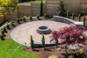 Round paver patio with fire-pit and retaining wall circled by plants.