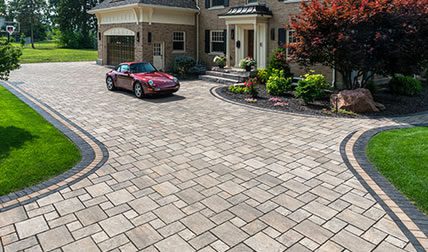 Driveway of a house made out of beige pavers with details in black. Red Porsche in the background.