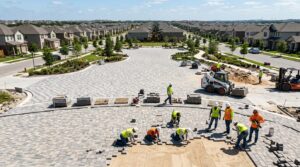 Wide shot of a construction site showing a large outdoor area being paved with grey and tan concrete pavers in sunlight.